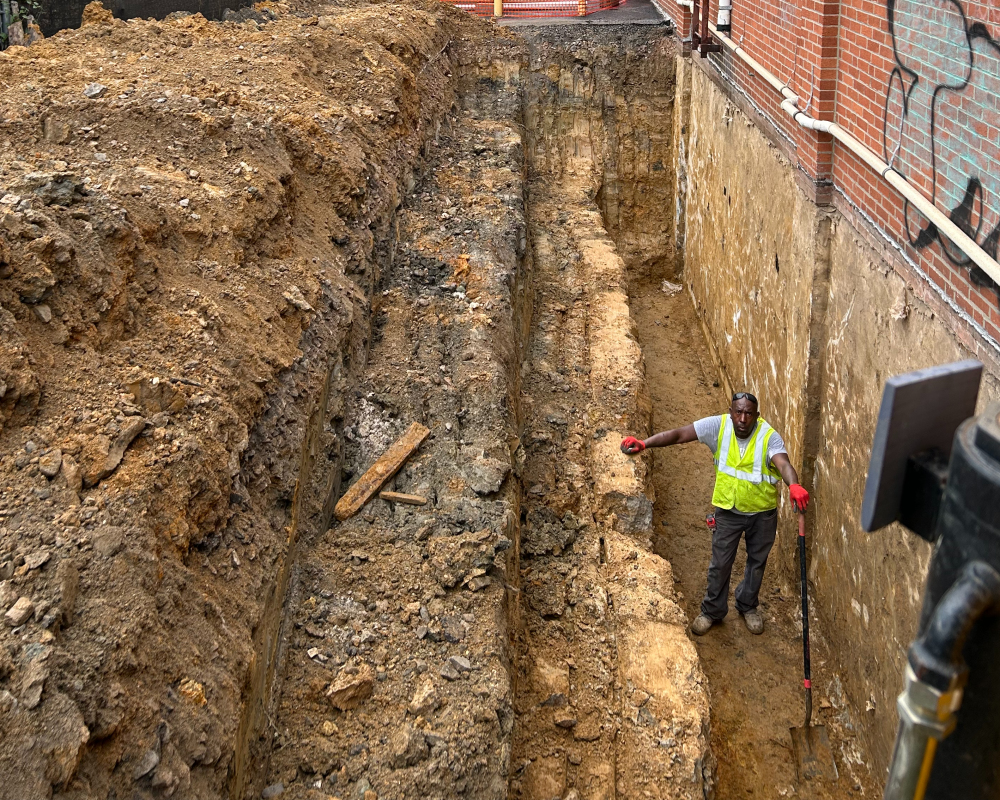 A worker stands in a deep trench beside a building foundation.